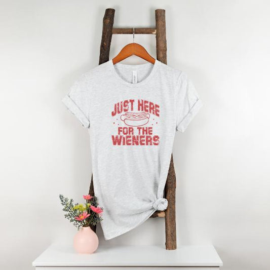 Just Here for the Wieners T-Shirt with red text and hot dog graphic on light gray fabric hanging on wooden ladder with pink vase and flowers
