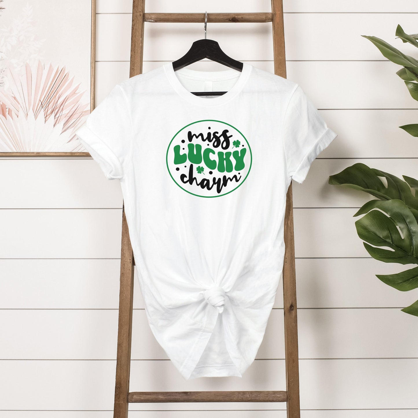 White Miss Lucky Charm T-shirt with green and black clover design hanging on wooden ladder against white shiplap wall