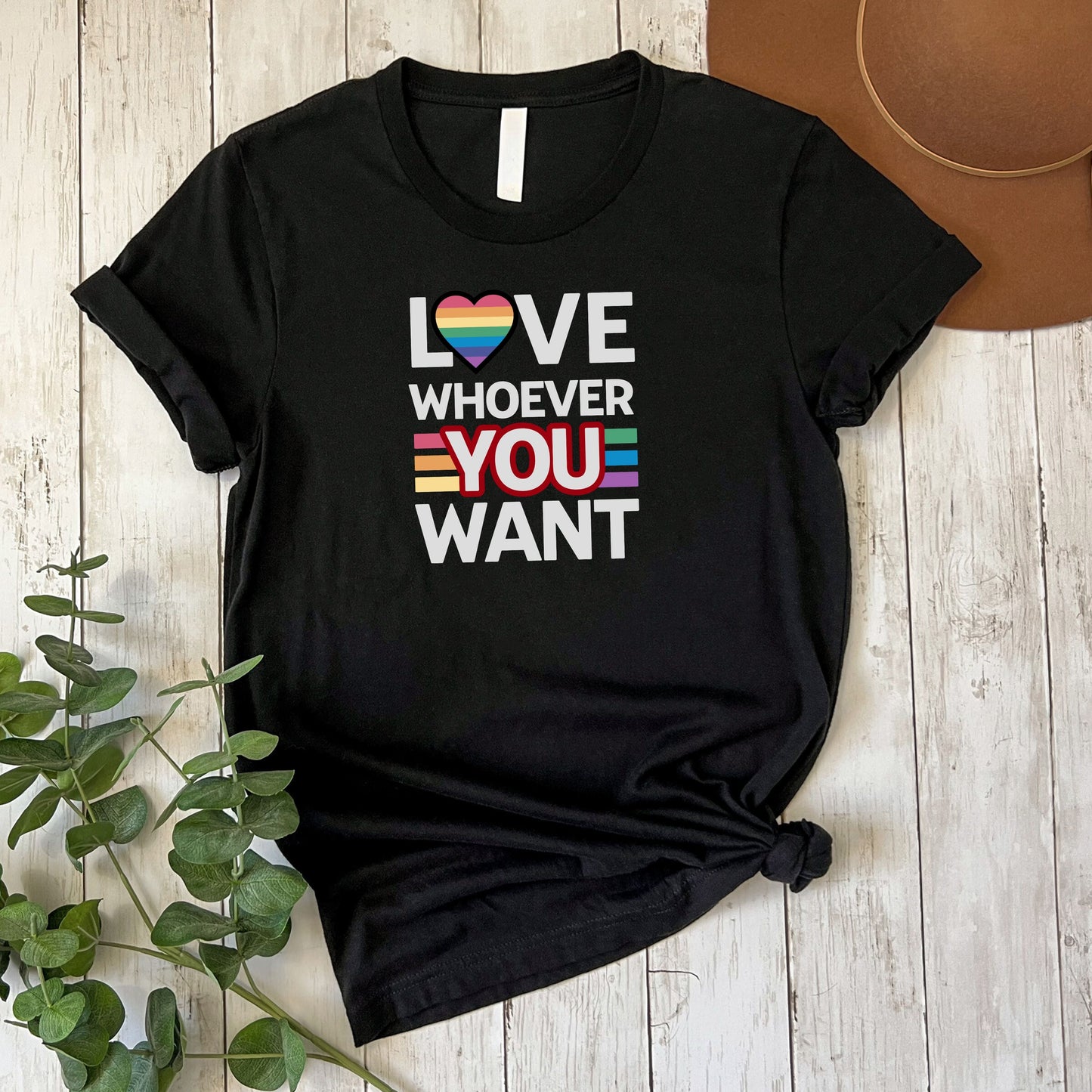 Black Love Whoever You Want T-shirt with rainbow heart and inclusive stripes design laid on wooden floor with green plant and brown hat nearby