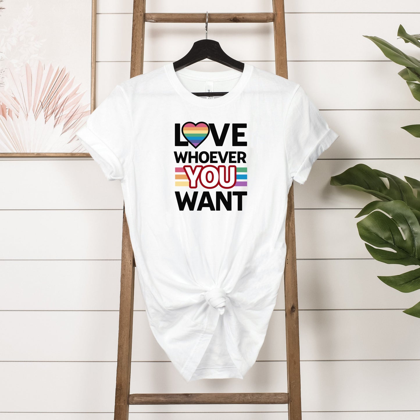 White Love Whoever You Want T-shirt with rainbow heart and inclusive pride colors hanging on wooden rack