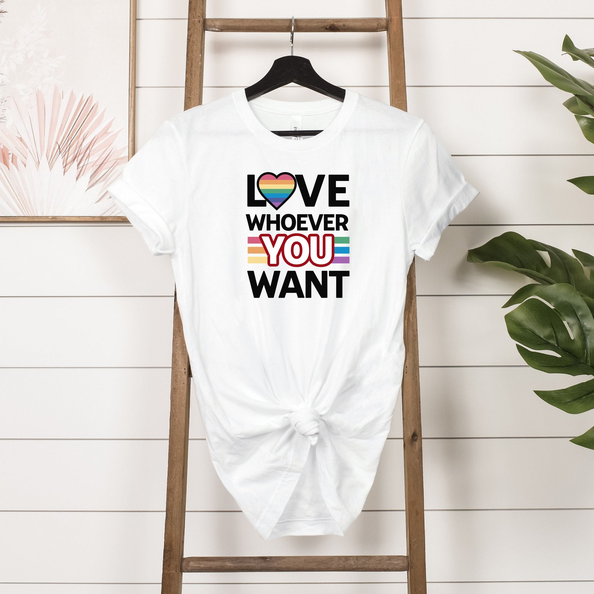 White Love Whoever You Want T-shirt with rainbow heart and inclusive pride colors hanging on wooden rack