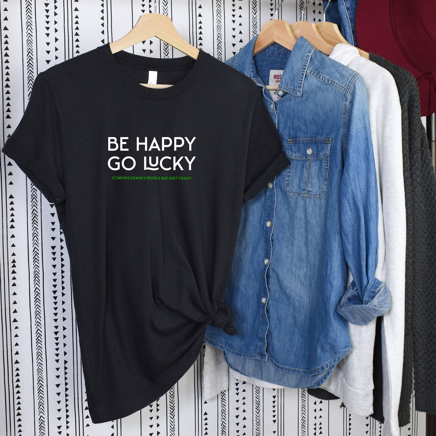Black Be Happy Go Lucky graphic t-shirt with knotted front detail on hanger next to denim and casual shirts.