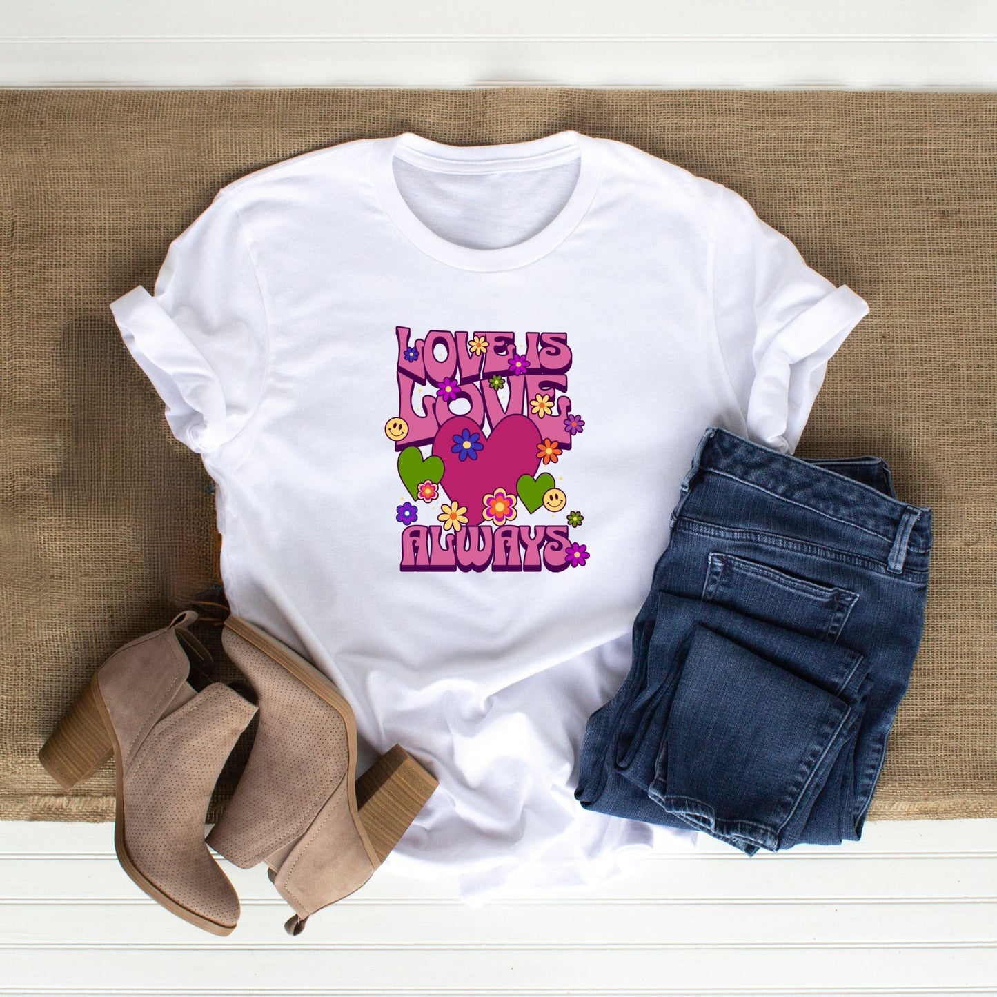 White "Love is Love Always" t-shirt with colorful heart and flower design, paired with blue jeans and beige heeled boots on a woven surface