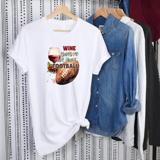 White t-shirt with "Wine Makes Me Care About Football" graphic featuring a glass of red wine and a football, displayed on a hanger with denim and gray shirts in the background